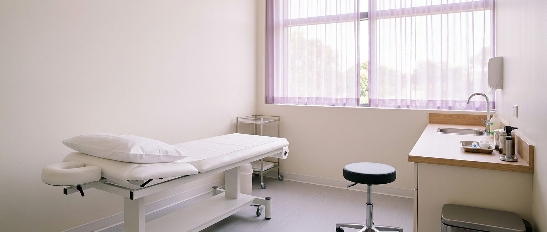 An unoccupied, softly lit medical exam room with a prepared exam table and a stool.